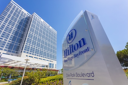 View from the ground looking up at a Hilton Hotel tower and sign against a clear blue sky.