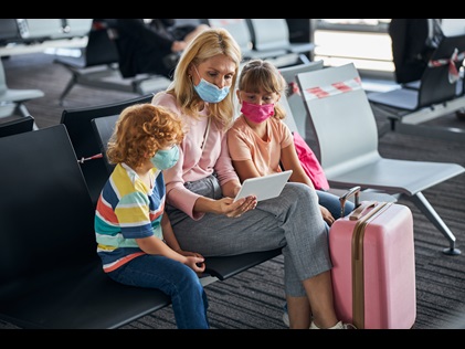 Mother will two small children wearing face coverings looking at a tablet at the airport. 