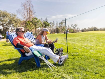 Two attendees relax on white lounge chairs along the side lines of the green at the Hilton Grand Vacations Tournament of Champions