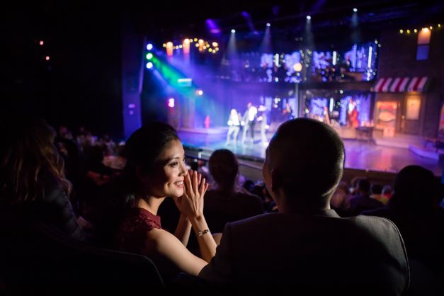 A couple sits in a theater, turned to each other, smiling, as the stage is lit up for a show, one of the experiences HGV Ultimate Access offers