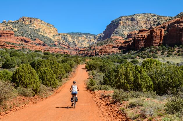 Wideshot image, woman riding bike along red dirt trail, stunning red rocks against blue sky, Sedona, Arizona. 