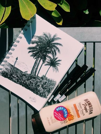 Reef safe sunscreen, pens and a sketchbook with a drawing of palm trees