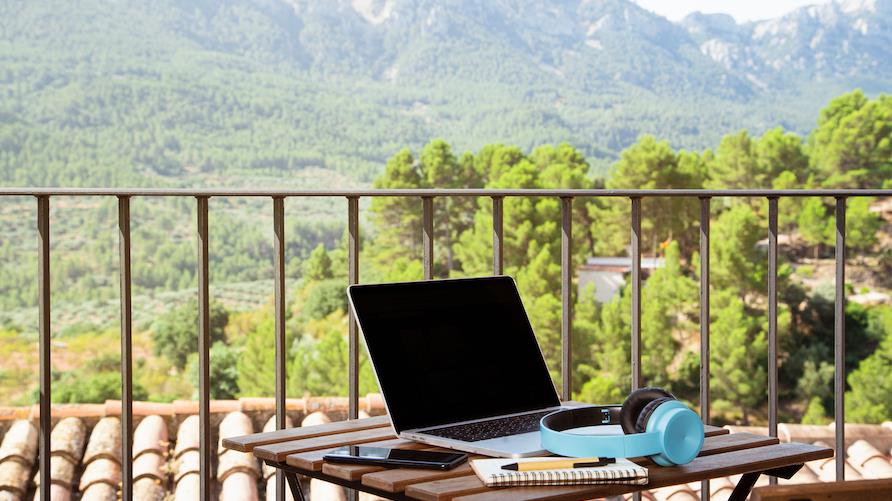 A laptop and blue headphones sit on a table on a balcony with a beautiful mountain view