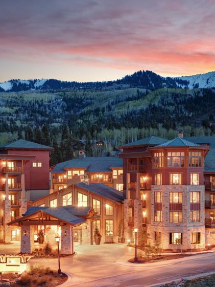 Exterior of Sunrise Lodge, a Hilton Grand Vacations Club in Park City, cozy lights on while the sun sets behind snowy mountains