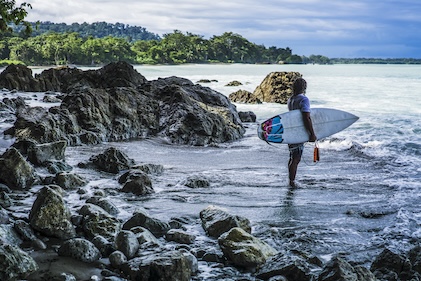 Lone surfer on rocky shore of the Osa Peninsula near Puerto Jiminez in Costa Rica. He is holding a surfboard as he watches the waves beyond.