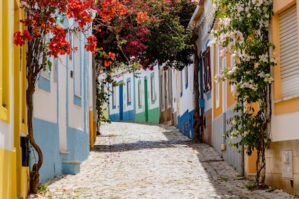 Colorful cobblestone streets of town on the Algarve coast featuring pastel homes, narrow roads lined with red and yellow bougainvillea.