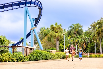 Family of four enjoys snacks as they walk through a theme park with large rollercoaster to the left of the frame.
