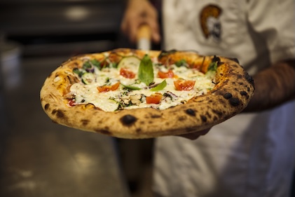 Close up of a chef holding a fire-roasted pizza and lots of toppings.