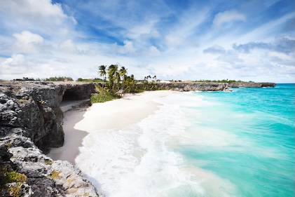 Turquoise blue waters at Barbados lap against the beach with black rock cliffs under a blue sky on a clear day.