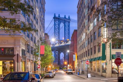 Urban street scene, small local shops, street lights glowing, dusk sky overhead, Brooklyn Bridge in distance, Dumbo Brooklyn, New York, New York. 