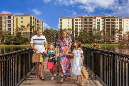 Young family walking along boardwalk, shopping bags, Tuscany Village, a Hilton Grand Vacations Club in distance, Orlando, Florida. 