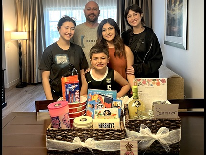 Young family of 5 poses facing the camera inside Marbrisa, a Hilton Grand Vacations Club with two very large gift baskets displaying treats, wine and gift cards.