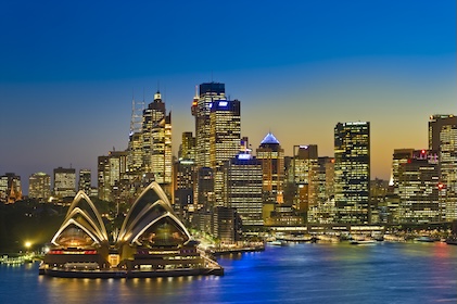 Tall buildings and the Sydney Opera house seen from the harbor at dusk. The lights in the skyscrapers and opera house cast a golden glow, and the remnants of the sunset are on the horizon.