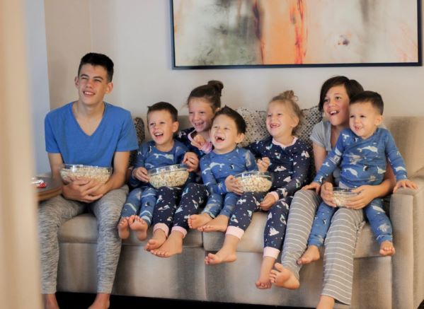 The seven Willis children on a couch, wearing pajamas, eating popcorn and laughing together while watching a movie