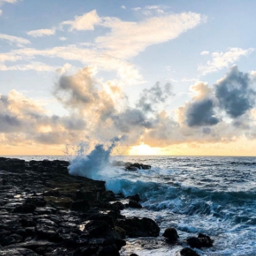 Foamy waves crash onto dark rocks on the Hawaiian beach at sunset.