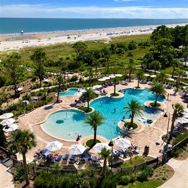 Overhead view of the bright blue pool and courtyard, ringed in palm trees with the South Carolina beach and Atlantic Ocean in the background.